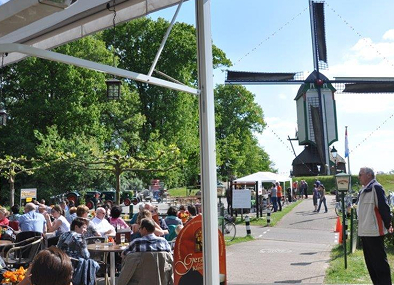 Outdoor waterfront cafe terrace with diners seated under umbrellas near historic windmill